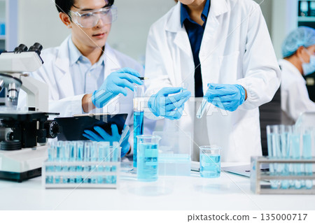 Closeup of Asian scientists experimenting with blue liquids and microscopes in a modern lab, representing biotechnology, chemistry, and medical innovation. 135000717