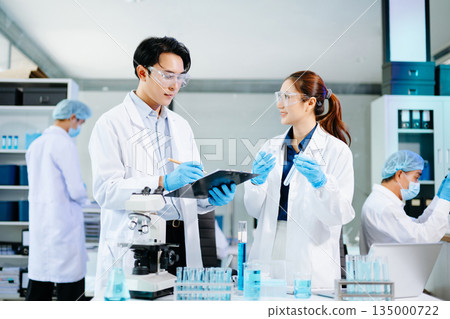 Scientists collaborating in a bright modern lab with blue liquids and microscopes, symbolizing teamwork, biotechnology, chemistry, and medical innovation. 135000722
