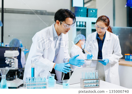 Scientists collaborating in a modern biotechnology lab with microscopes and test tubes, symbolizing teamwork, chemistry, and scientific innovation. 135000740