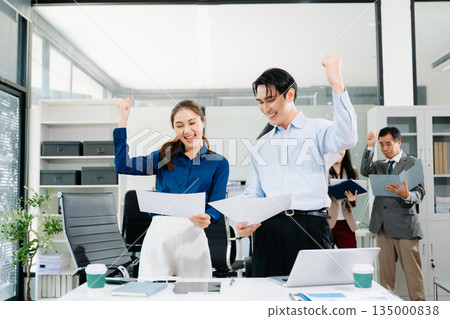 Joyful Asian business team celebrating success in a modern office, symbolizing teamwork, motivation, and achievement in corporate culture. 135000838