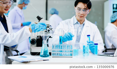 cientists working together in a modern lab with microscopes and blue liquids, symbolizing chemistry, biotechnology, and scientific innovation. 135000976