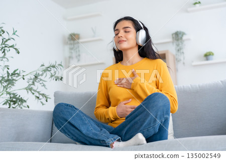 Happy young Asian woman practicing yoga and meditation at home sitting on couch Happy young Asian woman practicing yoga and meditation at home sitting on couch 135002549