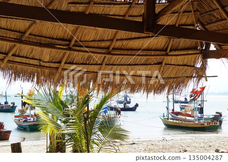 View from a bamboo house with a thatched roof to wooden fishing boats moored on the sea.View from bamboo house and thatched roof. View from a bamboo house with a thatched roof to wooden fishing boats moored on the sea.View from bamboo house and thatched roof. 135004287