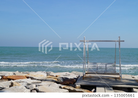 A beautiful place on the stone seashore with a swing against the backdrop of a blue sky and turquoise sea.A cozy place on the seashore. 135005661