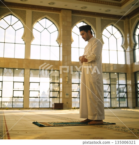 Muslim man performing Salah prayer inside a mosque. Side view of a male in white thobe standing on a prayer mat. Islamic religion and Ramadan concept 135006321