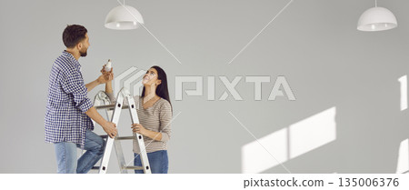 Young man standing on the ladder changing light bulb in the room with his smiling wife. Young man standing on the ladder changing light bulb in the room with his smiling wife. 135006376