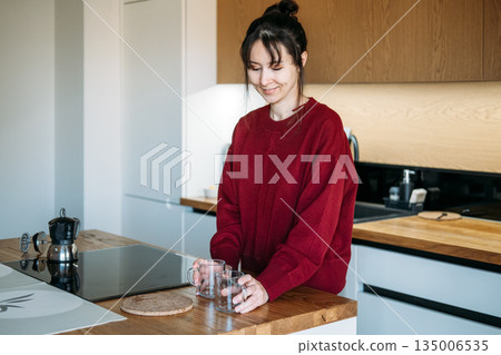 Woman places glass mugs on wooden counter near moka pot in kitchen. Artisanal coffee preparation, home brewing techniques, third-wave coffee culture. 135006535
