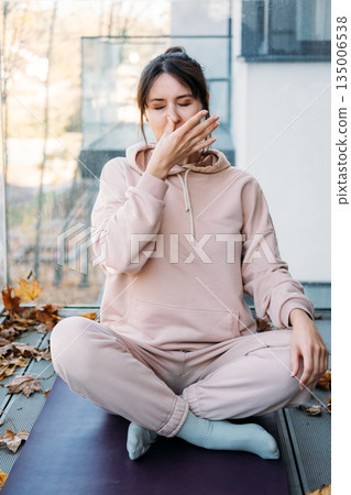 Woman practices Nadi Shodhana pranayama while sitting on yoga mat on balcony with autumn leaves. Mindfulness, outdoor wellness, mental health. Woman practices Nadi Shodhana pranayama while sitting on yoga mat on balcony with autumn leaves. Mindfulness, outdoor wellness, mental health. 135006538