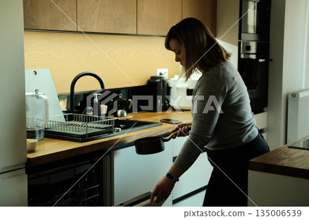 Woman unloads dishwasher in modern kitchen while holding small saucepan. Kitchen organization, domestic labor, home management, minimalist living Woman unloads dishwasher in modern kitchen while holding small saucepan. Kitchen organization, domestic labor, home management, minimalist living 135006539