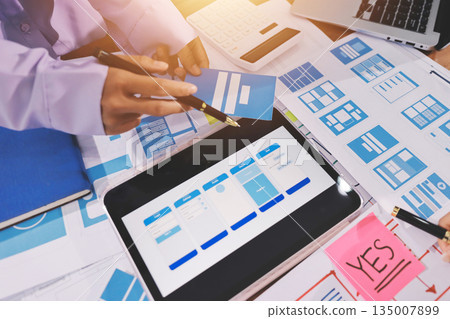 Close up ux developer and ui designer brainstorming about mobile app interface wireframe design on table with customer breif and color code at modern office.Creative digital development agency. 135007899