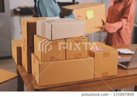 Startup SME small business entrepreneur of freelance Asian woman wearing apron using laptop and box to receive and review orders online to prepare to pack sell to customers, online sme business ideas. Startup SME small business entrepreneur of freelance Asian woman wearing apron using laptop and box to receive and review orders online to prepare to pack sell to customers, online sme business ideas. 135007909