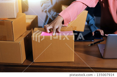 Startup SME small business entrepreneur of freelance Asian woman wearing apron using laptop and box to receive and review orders online to prepare to pack sell to customers, online sme business ideas. Startup SME small business entrepreneur of freelance Asian woman wearing apron using laptop and box to receive and review orders online to prepare to pack sell to customers, online sme business ideas. 135007914