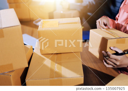 Startup SME small business entrepreneur of freelance Asian woman wearing apron using laptop and box to receive and review orders online to prepare to pack sell to customers, online sme business ideas. 135007925