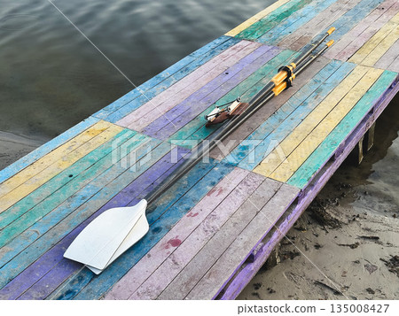Colorful dock with oar by calm river, perfect for boating adventures. Colorful dock with oar by calm river, perfect for boating adventures. 135008427