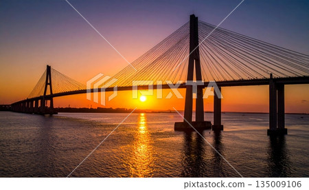 A vibrant sunset and sunrise reflect over the city river and calm lake water near the bridge and harbor pier as clouds fill the summer sky at dusk 135009106