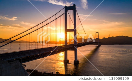A vibrant sunset and sunrise reflect over the city river and calm lake water near the bridge and harbor pier as clouds fill the summer sky at dusk A vibrant sunset and sunrise reflect over the city river and calm lake water near the bridge and harbor pier as clouds fill the summer sky at dusk 135009109
