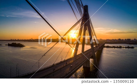 A vibrant sunset and sunrise reflect over the city river and calm lake water near the bridge and harbor pier as clouds fill the summer sky at dusk A vibrant sunset and sunrise reflect over the city river and calm lake water near the bridge and harbor pier as clouds fill the summer sky at dusk 135009110