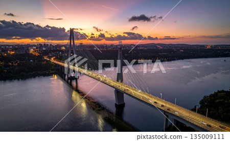 A vibrant sunset and sunrise reflect over the city river and calm lake water near the bridge and harbor pier as clouds fill the summer sky at dusk 135009111