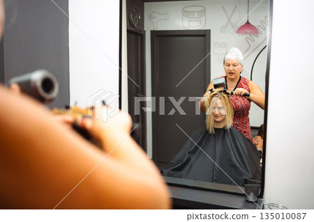 Hairdresser styling woman's hair in salon, reflecting in mirror 135010087