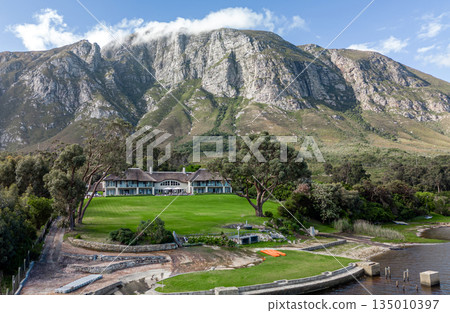 Residential building nestled at foot of large mountain with green lawn near lake Residential building nestled at foot of large mountain with green lawn near lake 135010397