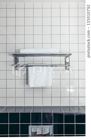 A clean bathroom with white ceramic tiles. A metal towel rack holds two neatly folded white towels. The floor features dark ceramic tiles. A clean bathroom with white ceramic tiles. A metal towel rack holds two neatly folded white towels. The floor features dark ceramic tiles. 135010792