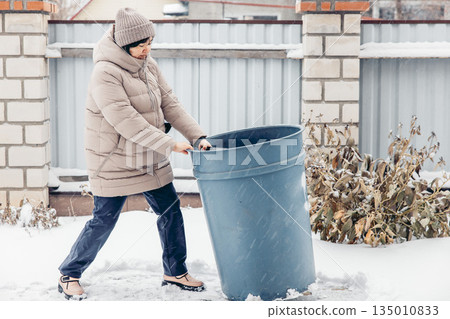 middle-aged Asian woman in a beige coat and hat pushes a large blue trash bin through the snow in a winter setting. 135010833