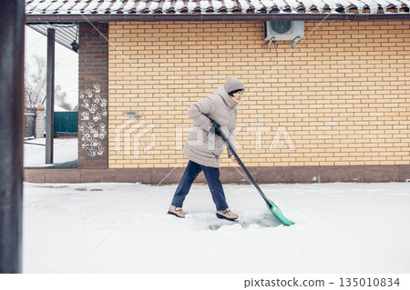 A young Asian woman in a beige winter coat shovels snow outside a brick house, while clearing the snow. A young Asian woman in a beige winter coat shovels snow outside a brick house, while clearing the snow. 135010834