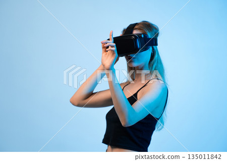 Young woman adjusting virtual reality headset with hand gesture, immersive VR experience and futuristic technology concept on blue background 135011842