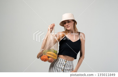 Eco conscious woman looking at fresh groceries in reusable net bag, sustainable food shopping and plastic free lifestyle concept 135011849