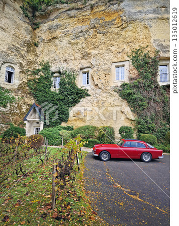 Rochecorbon, France - October 24, 2025: A vintage red sports car parked near the limestone cliffs of Les Hautes Roches. Dwellings are carved directly into the golden rock, draped in lush green ivy 135012619