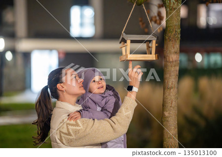 Mother holding daughter and pointing at bird feeder at night outdoors. Shared discovery and learning moment during walk. Mother holding daughter and pointing at bird feeder at night outdoors. Shared discovery and learning moment during walk. 135013100