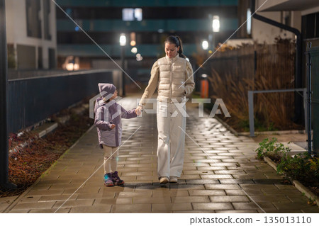 Mother and child walking hand in hand through wet street at night. Rainy parenting scene. Mother and child walking hand in hand through wet street at night. Rainy parenting scene. 135013110