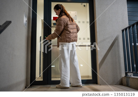 Woman unlocking entrance door of apartment building at night. Returning home after day out. Woman unlocking entrance door of apartment building at night. Returning home after day out. 135013119