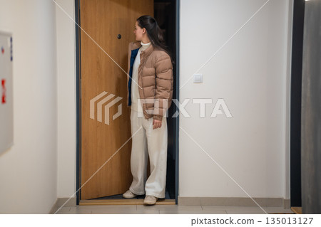 Woman standing in corridor near apartment door before leaving home. Daily transition between home and city life. 135013127
