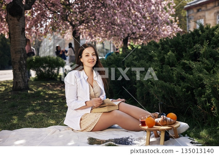 spring scene, picnic setup, seasonal fruit. Sitting on picnic blanket under blooming cherry trees, young woman enjoys book and holds flower, with snacks and fruit nearby. spring scene, picnic setup, seasonal fruit. Sitting on picnic blanket under blooming cherry trees, young woman enjoys book and holds flower, with snacks and fruit nearby. 135013140