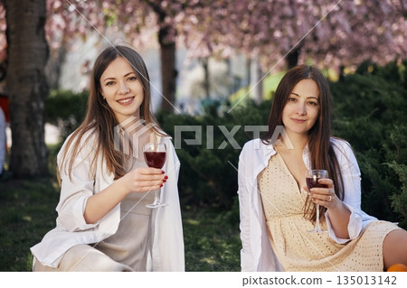 Smiling Women, Blooming Flowers, Friends Laughing. Two young women are smiling and holding wine glasses while relaxing in lush spring garden with blooming trees. Smiling Women, Blooming Flowers, Friends Laughing. Two young women are smiling and holding wine glasses while relaxing in lush spring garden with blooming trees. 135013142