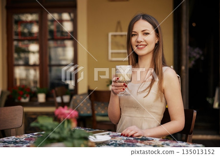 sidewalk cafe, Relaxed Setting, Pleasant Atmosphere. At outdoor cafe, young woman smiles while holding coffee cup, creating warm and relaxed atmosphere. sidewalk cafe, Relaxed Setting, Pleasant Atmosphere. At outdoor cafe, young woman smiles while holding coffee cup, creating warm and relaxed atmosphere. 135013152