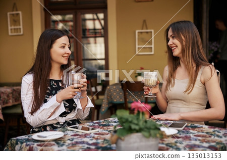 enjoying coffee, Coffee Break, Friendly Atmosphere. Two young women engage in relaxed coffee conversation at open-air cafe with warm tones. 135013153