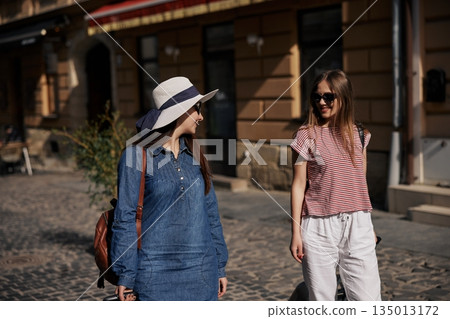 walking down street, happy travelers, street travel. Carrying suitcases, two casually dressed young women walk down cobblestone street enjoying sunny urban atmosphere. 135013172