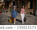 city street, travel companions, urban exploration. Two young women in casual attire stroll down cobblestone street with suitcases on sunny day in city. 135013173