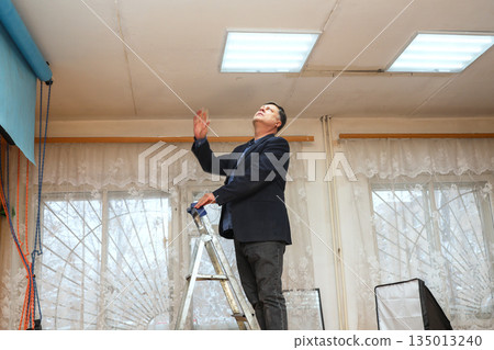 Man on Step Ladder in Photo Studio Checking Ceiling Light Fixture Man on Step Ladder in Photo Studio Checking Ceiling Light Fixture 135013240