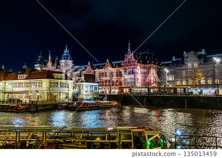 Amsterdam Railway station canal water house building. Netherlands night. Evening street architecture. Water boats Amsterdam Railway station canal water house building. Netherlands night. Evening street architecture. Water boats 135013459