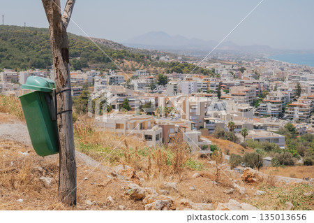 Green plastic trash bin or garbage can attached to a tree on a dry hill overlooking a coastal mediterranean town on a hot sunny day Green plastic trash bin or garbage can attached to a tree on a dry hill overlooking a coastal mediterranean town on a hot sunny day 135013656