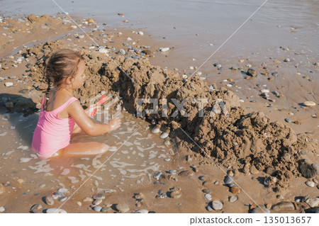Little girl in a pink swimsuit playing in the small waves and wet sand, building a sand fort on a pebble beach during a sunny summer day 135013657