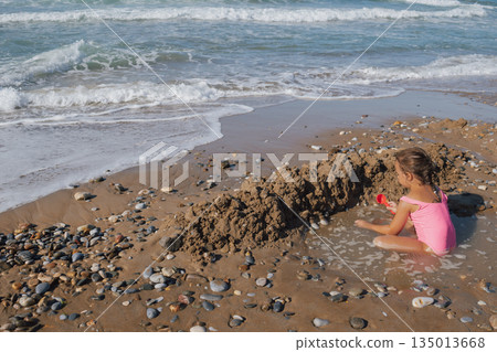 Happy Little Girl in Pink Swimsuit Building Sand Castle Moat on a Pebble Beach with Waves and Sea Foam in Summer Sunlight 135013668