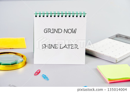 A notepad displaying the message Grind Now Shine Later sits among office supplies such as paper clips, sticky notes, calculator, and a magnifying glass on a smooth surface 135014004