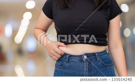 Young woman in black crop top and blue jeans, standing confidently in a bright indoor space, showcasing body positivity and self-acceptance in a modern environment 135014240