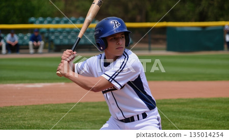 Young male baseball player in white uniform and blue helmet stands ready to bat on a sunny day at the baseball field, showcasing athleticism and focus in a competitive environment 135014254