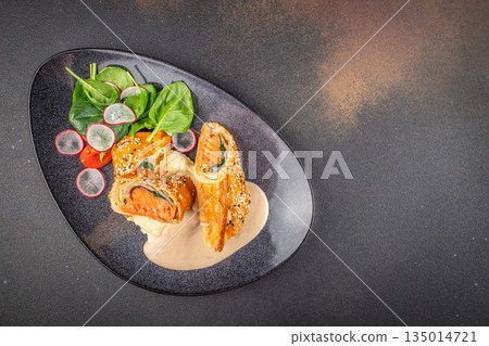 Salmon wrapped in puff pastry with creamy sauce and fresh salad served on dark plate with copy space Salmon wrapped in puff pastry with creamy sauce and fresh salad served on dark plate with copy space 135014721