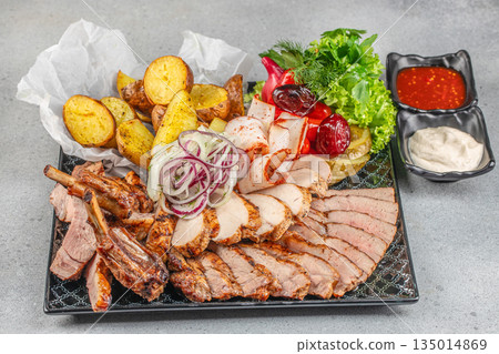 Grilled meat platter with baked potatoes, vegetables, and sauces on a black plate 135014869